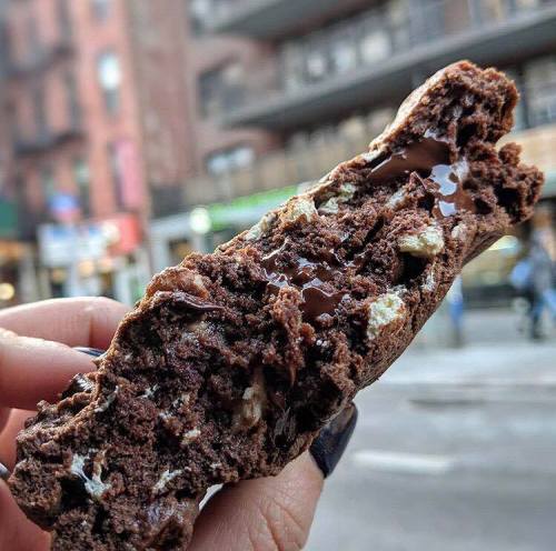 A hand holds a gooey chocolate chip cookie, revealing its soft, melted interior against a city backdrop.