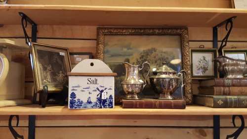 A wooden shelf displaying various antiques, including a salt box, silver pitchers, framed art, and old books.