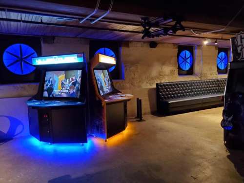 Two arcade machines with colorful lights in a dimly lit room, featuring a black leather couch and circular windows.
