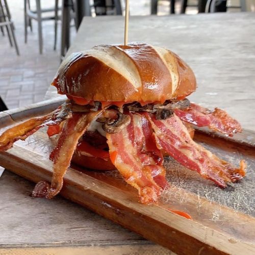 A pretzel bun burger overflowing with crispy bacon and toppings, served on a wooden board.