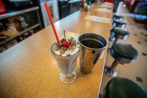 A chocolate milkshake topped with whipped cream, a cherry, and chocolate shavings, served with a metal shaker.