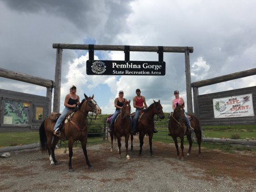 Four riders on horseback pose in front of the Pembina Gorge State Recreation Area sign under a cloudy sky.