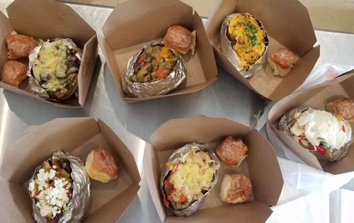 Six takeout boxes containing baked potatoes topped with various ingredients, accompanied by small rolls.