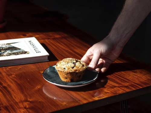 A hand reaches for a muffin on a black plate, with a book titled "Nashville" in the background on a wooden table.