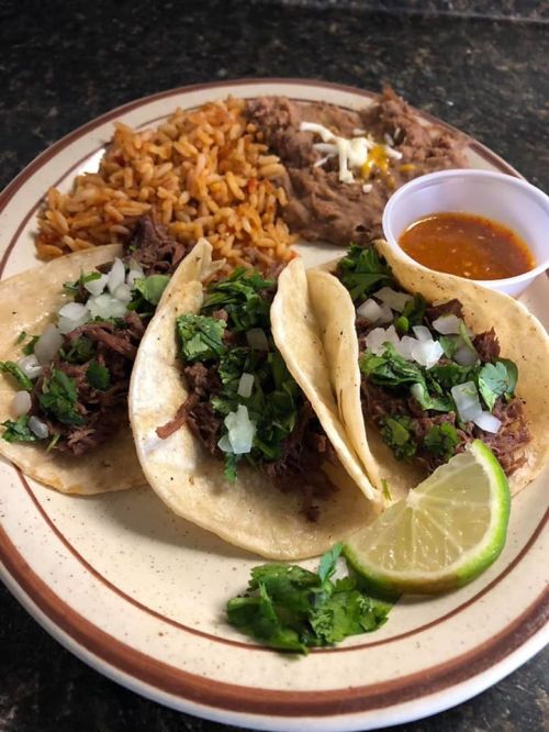 A plate with three tacos filled with beef, topped with onions and cilantro, served with rice, beans, and a lime wedge.