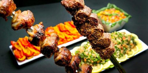 A skewer of grilled meat in the foreground, with colorful sides of vegetables and salads in the background.