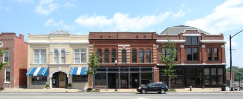 Historic buildings line a street, featuring varied architectural styles and storefronts under a blue sky.