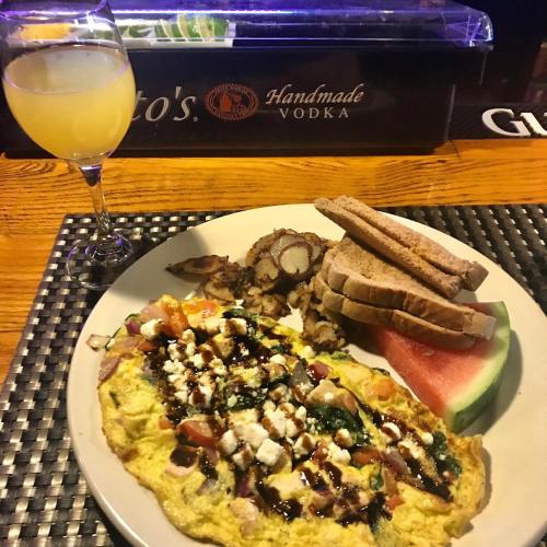 A plate with a colorful omelette, toast, potatoes, and watermelon, alongside a glass of mimosa on a bar counter.