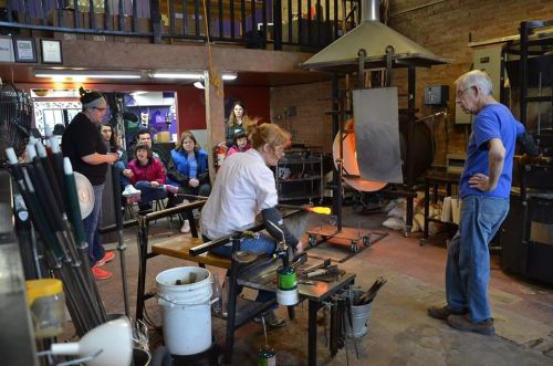 A glassblowing demonstration in a workshop, with an artist working and an audience observing. Tools and equipment are visible.