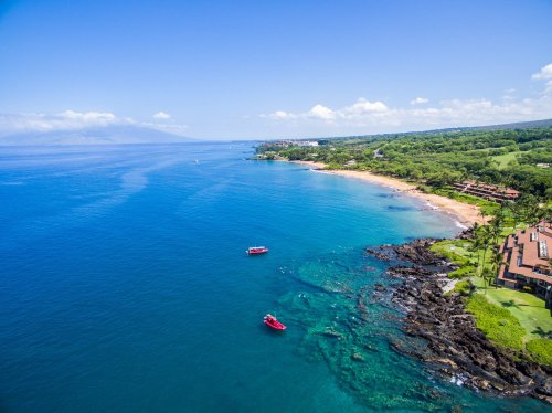 Aerial view of a tropical coastline with clear blue water, sandy beach, and boats near lush green hills.