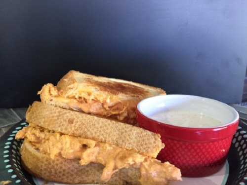 A plate with two grilled cheese sandwiches filled with orange chicken and a small bowl of dipping sauce.