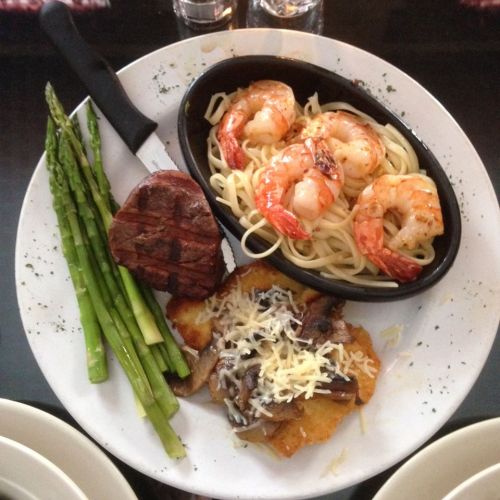 A plate featuring grilled shrimp over pasta, a steak, sautéed mushrooms, and asparagus.