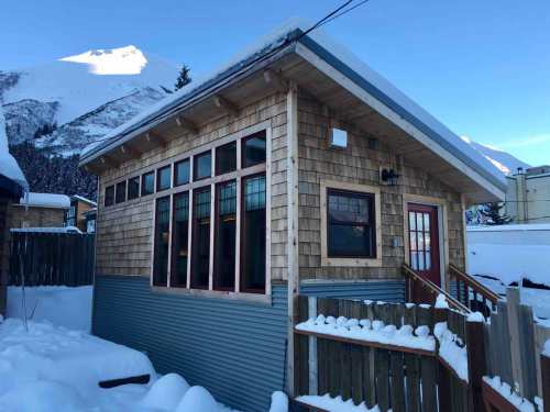 A cozy wooden cabin with large windows, surrounded by snow, against a backdrop of mountains and a clear blue sky.