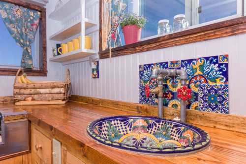 Colorful tiled sink with a wooden countertop, surrounded by rustic decor and bright window curtains.