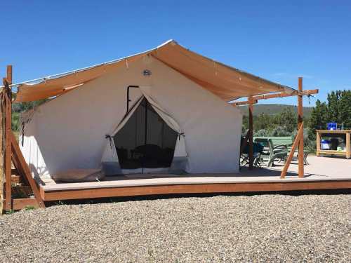 A spacious white tent on a wooden deck, surrounded by gravel and greenery, under a clear blue sky.