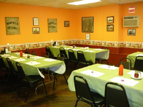 A cozy dining room with green tablecloths, black chairs, and vintage decor on orange walls.