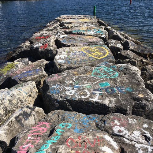 A rocky pier covered in colorful graffiti, extending into a calm body of water under a clear blue sky.
