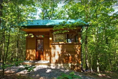 A small wooden cabin with a green roof, surrounded by lush trees in a forested area.