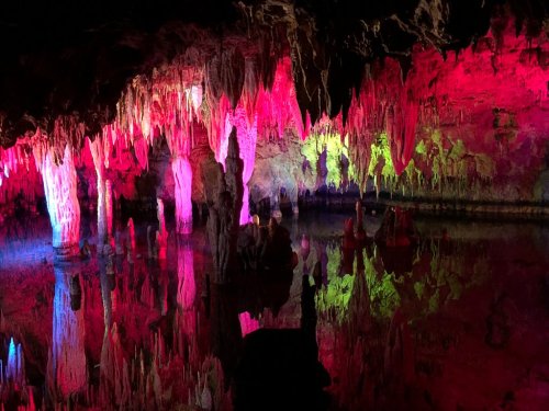 Colorful stalactites and stalagmites illuminated in a cave, reflecting in still water below.