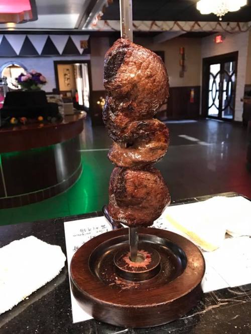 A skewer with three pieces of grilled meat, resting on a wooden base in a restaurant setting.