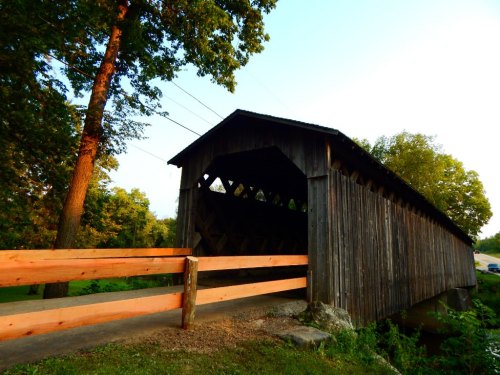 A rustic wooden covered bridge with a stone foundation, surrounded by trees and a clear sky.