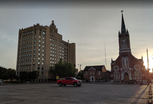 A red truck parked in a lot beside a tall building and a historic church at sunset.