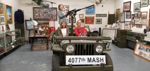 Three women pose in a military jeep inside a museum, with various historical artifacts displayed in the background.
