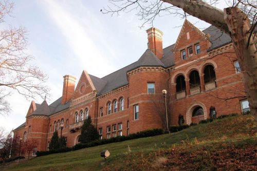 Historic brick building with ornate architecture, large windows, and a sloped lawn in front, under a clear sky.
