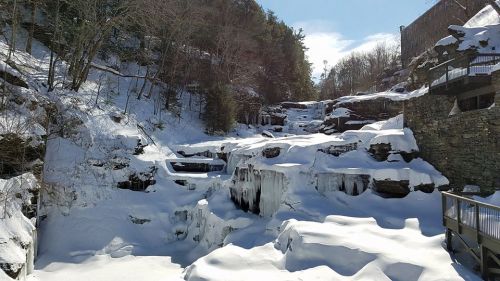 A snowy landscape with frozen waterfalls and rocky cliffs, surrounded by trees in a winter setting.