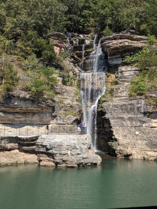 A serene waterfall cascading over rocky cliffs into a calm green pool, with a person standing on a nearby rock.