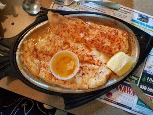 A sizzling platter of fish fillets with a side of melted butter and a lemon wedge, served on a restaurant table.