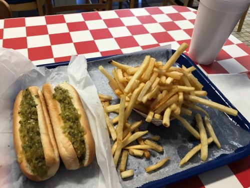 Two hot dogs with green relish on a tray, accompanied by a large serving of French fries and a drink.