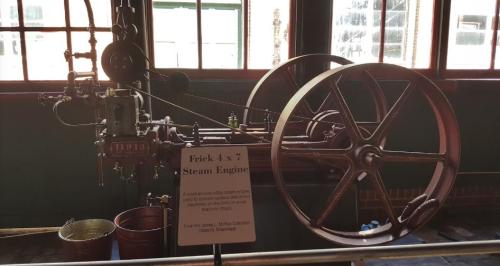 A vintage Erick 4x7 steam engine displayed indoors, with large wheels and a sign detailing its history.