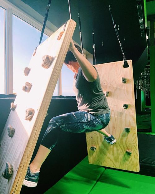 A person climbs a wooden wall with rock holds, suspended in a gym with large windows and green padding below.