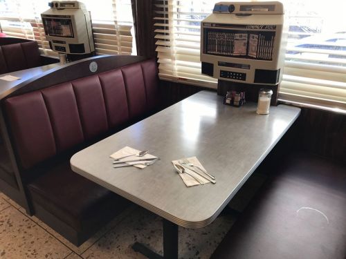 A diner booth with a retro jukebox, set with utensils and napkins on a shiny table. Sunlight filters through the blinds.