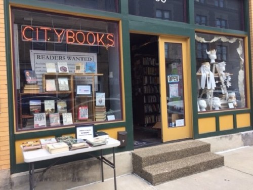 A cozy bookstore with a neon "CITY BOOKS" sign, open door, and a table displaying books outside.