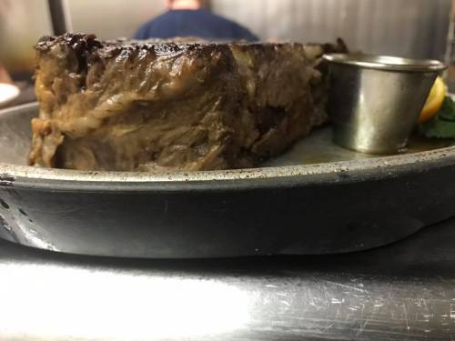 A close-up of a juicy, grilled steak on a metal plate, accompanied by a small cup and a slice of lemon.