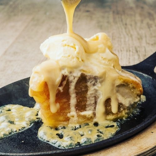 A slice of warm dessert topped with melting ice cream and drizzled with sauce, served on a sizzling skillet.