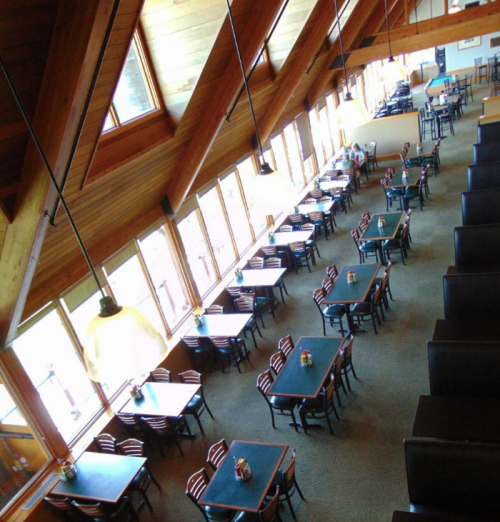 A spacious restaurant interior with wooden beams, rows of tables, and large windows letting in natural light.
