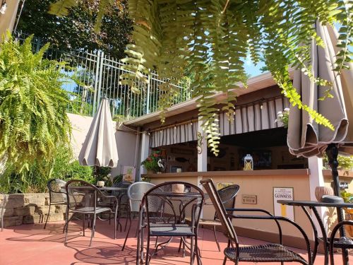 A cozy outdoor seating area with black chairs, umbrellas, and lush greenery near a bar.