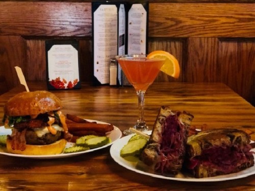 A burger with fries and a cocktail next to a sandwich with pickles on a wooden table.