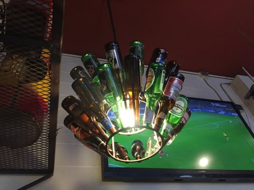 A unique chandelier made from various beer bottles, illuminated from the center, with a TV showing a soccer game in the background.