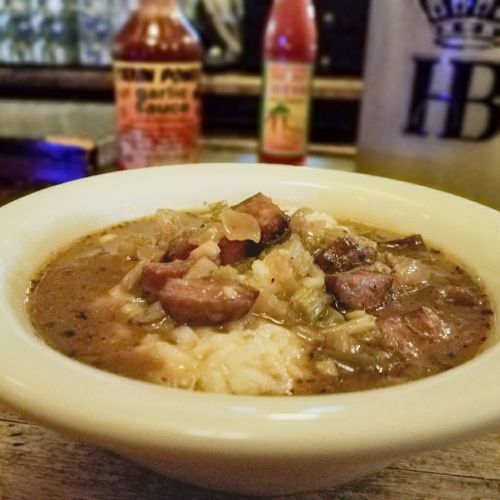 A bowl of gumbo with sausage and vegetables served over rice, with hot sauce bottles in the background.