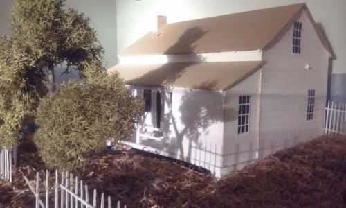 A miniature white house with a brown roof, surrounded by a fence and a tree, set on a bed of straw.