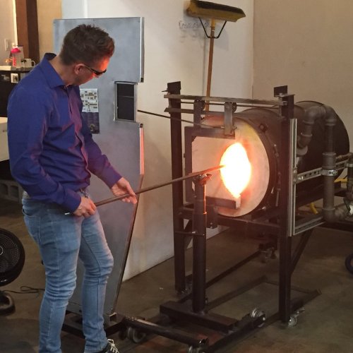 A person in a blue shirt uses a blowpipe to shape molten glass in a glassblowing studio.