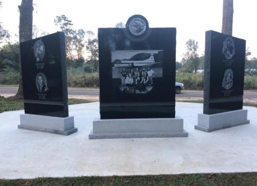 A memorial with three black granite panels featuring engraved images and text, set in a natural outdoor setting.