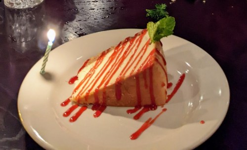 A slice of cheesecake drizzled with red sauce, topped with a mint leaf and a lit birthday candle on a white plate.