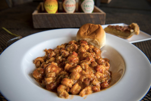 A plate of shrimp étouffée with a soft bun, accompanied by hot sauce bottles in the background.