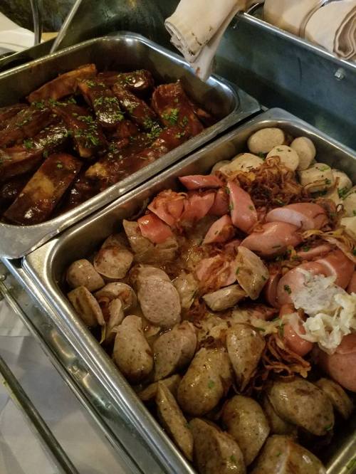 Two trays of food: one with glazed ribs and the other with assorted sausages, potatoes, and sauerkraut, garnished with herbs.