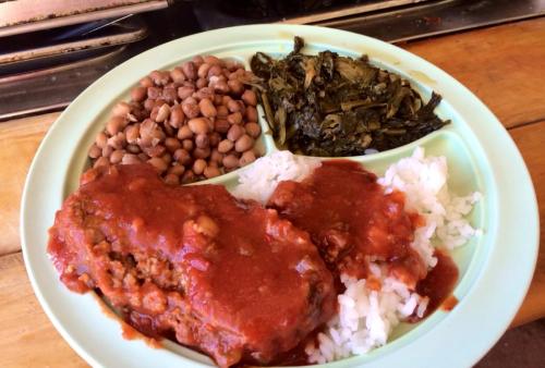 A plate of food featuring meatloaf with sauce, rice, collard greens, and a side of pinto beans.